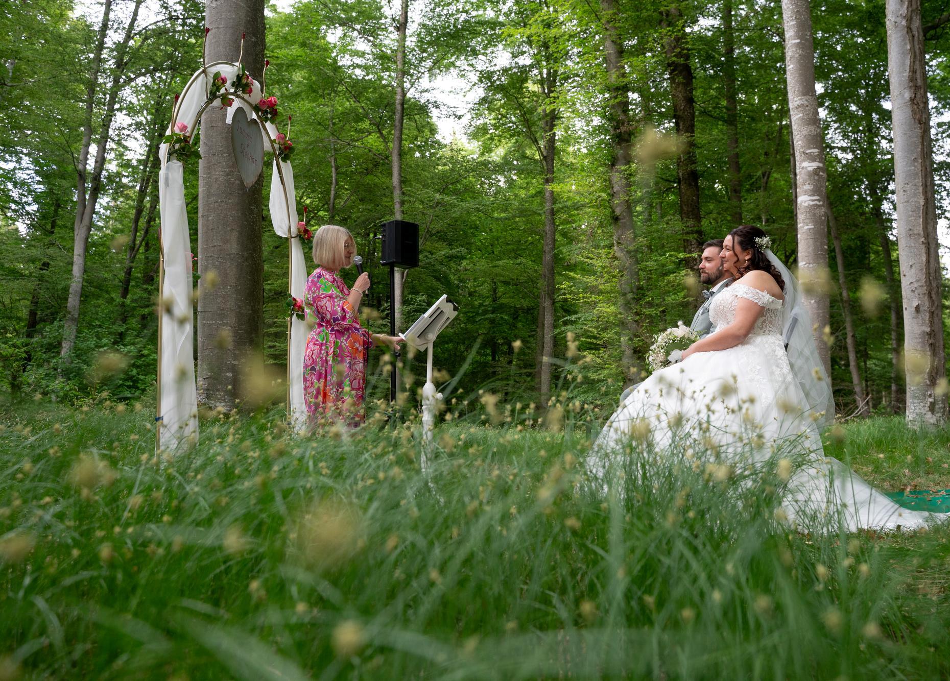 Ein Hochzeitspaar sitzt nebeneinander im Wald und schaut auf eine freie Rednerin. Diese steht vor ihnen mit einem Mikrophon in der Hand unter einem Traubogen. Im Vordergrund ist viel Gras und im Hintergrund güne Bäume zu sehen.