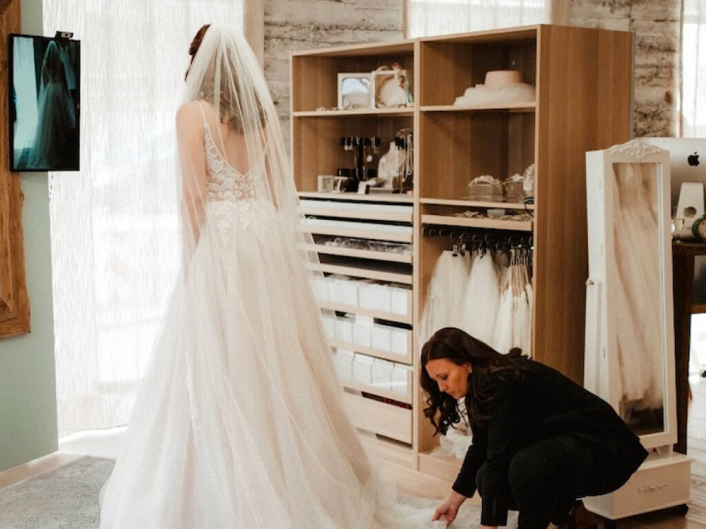 Eine Frau in einem eleganten Hochzeitskleid steht mit dem Rücken zur Kamera in einem Raum mit Holzmöbeln und Brautmodenartikeln. Hinter ihr kniet eine weitere Frau in einem schwarzen Anzug und richtet sorgfältig den langen Schleier.