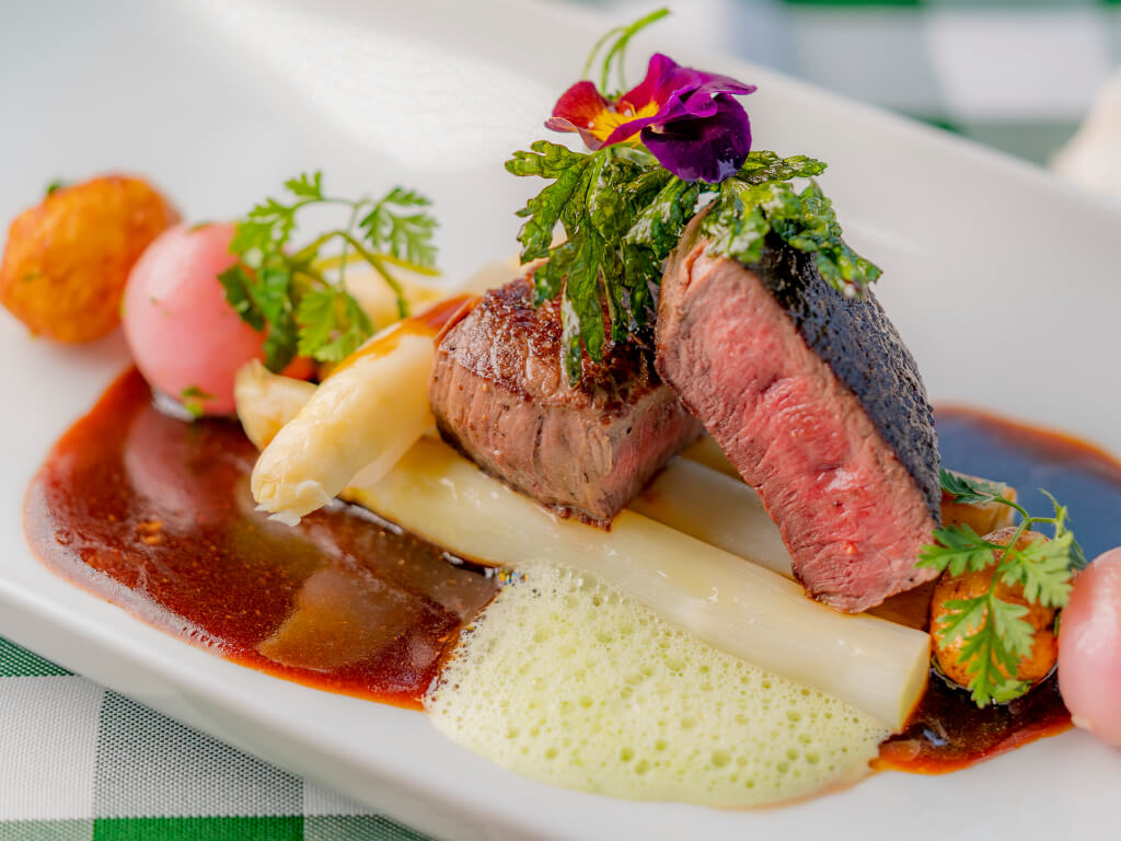 Eine Nahaufnahme eines stilvoll angerichteten Tellers mit einem perfekt gebratenen Steak, Spargel und verschiedenen frischen Kräutern in einer aromatischen Soße. Die Farben und Texturen der Zutaten heben die feine Komposition des Gerichts hervor.
