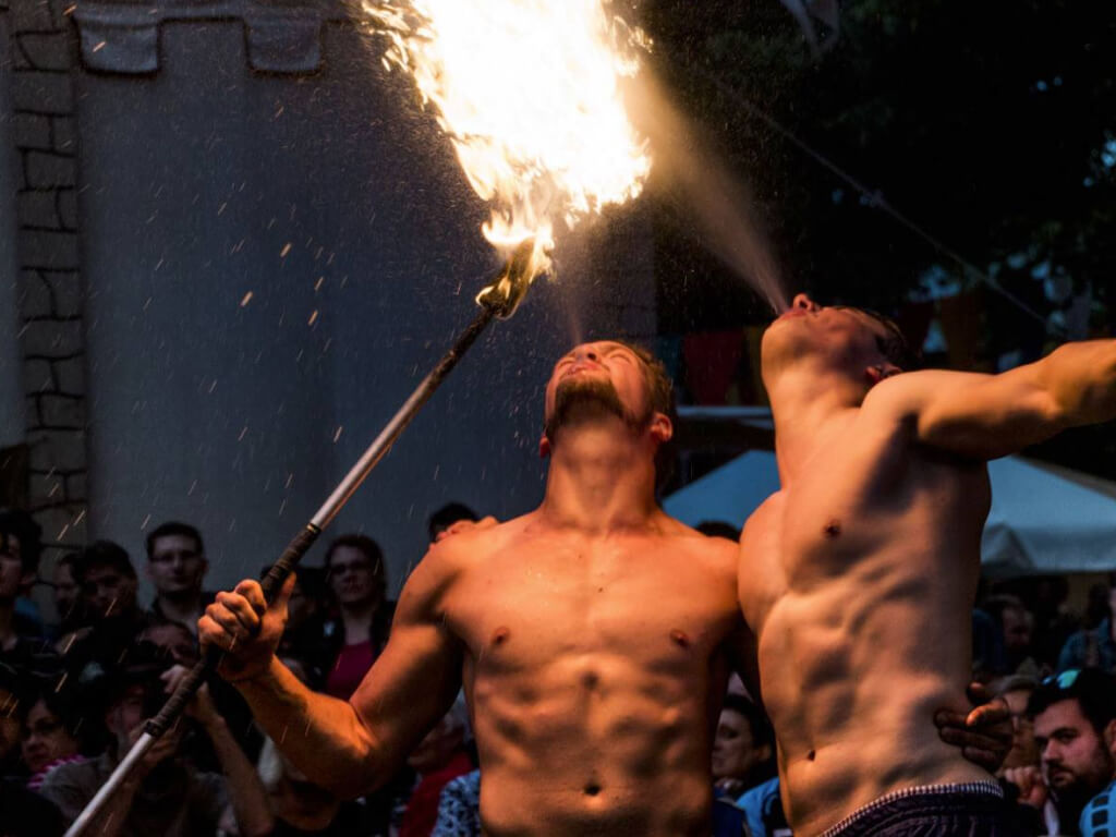 Zwei oberkörperfreie Feuerkünstler führen eine spektakuläre Feuerspuck-Show vor einem faszinierten Publikum auf. Einer der Künstler hält eine lange Fackel mit brennender Flamme, während der andere eine riesige Feuerwolke in die Luft bläst. Die Szene spielt sich in einer stimmungsvollen, abendlichen Kulisse mit mittelalterlich anmutenden Dekorationen und bunten Wimpeln im Hintergrund ab.