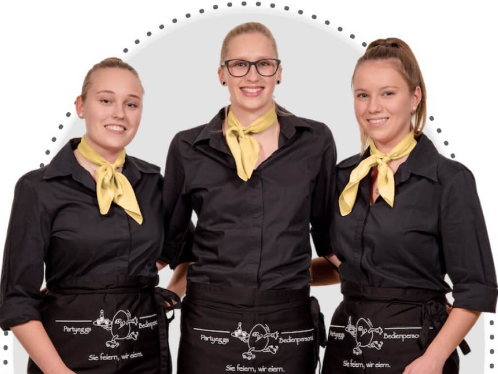 Drei Frauen stehen nebeneinander, jeweils in einer schwarzen Bluse und einer schwarzen Schürze gekleidet. Um ihren Hals tragen sie ein gelbes Tuch, das einen lebendigen Farbkontrast zu ihrer dunklen Kleidung bildet. Ihre Haltung wirkt harmonisch und kooperativ, was eine einladende und freundliche Atmosphäre ausstrahlt.