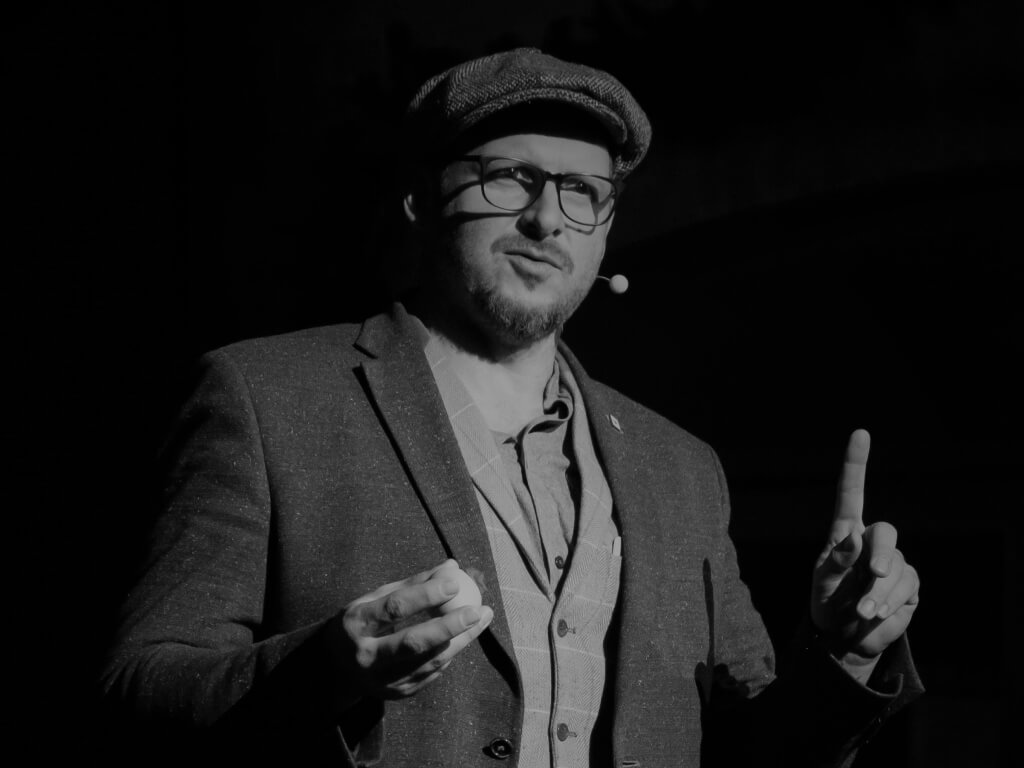 Das Schwarz-Weiß-Bild zeigt einen Mann im Anzug, der einen Gegenstand in der einen Hand hält, während er mit dem erhobenen Zeigefinger eine Geste macht. Er trägt eine Brille, ein Headset und eine Mütze, was ihm einen modernen und zugleich professionellen Look verleiht.