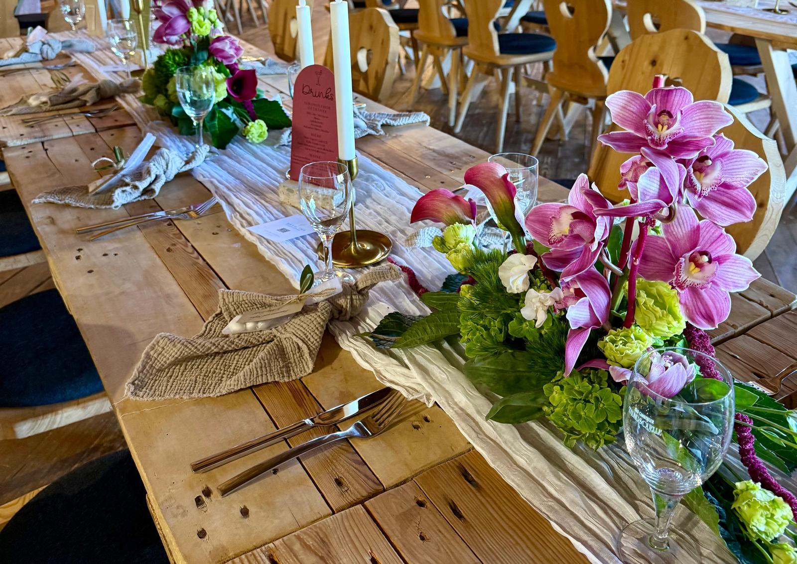 Ein gedeckter Tisch mit lila Orchideen in einem grünen Gesteck. In der Mitte liegt ein weißem Tischläufer mit Stabkerzen.